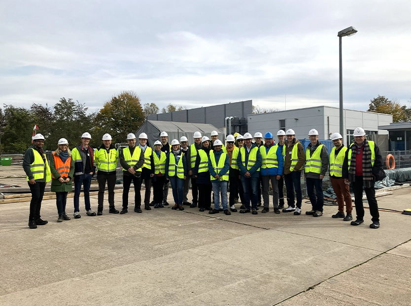 Eine Gruppe von etwa 20 Personen in Sicherheitswesten und weißen Schutzhelmen steht auf einem Industriegelände vor einem grauen Gebäude. Im Hintergrund sind Bäume und Baustellenmaterial zu sehen.