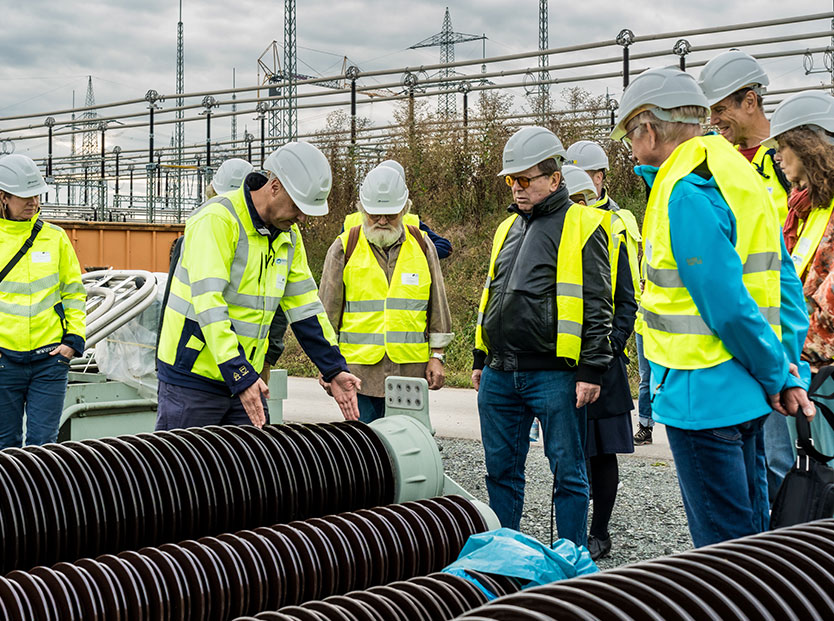 Das Bild zeigt eine Gruppe von Menschen in gelben Warnwesten und Bauhelmen bei einer Führung im Umspannwerk. Ein Mann erklärt einen Isolator und misst ihn mit seinen Händen ab. Die anderen Teilnehmenden stehen um ihn herum und hören aufmerksam zu. Im Hintergrund sind die Strommasten und die technische Infrastruktur des Umspannwerks zu sehen.