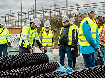 Das Bild zeigt eine Gruppe von Menschen in gelben Warnwesten und Bauhelmen bei einer Führung im Umspannwerk. Ein Mann erklärt einen Isolator und misst ihn mit seinen Händen ab. Die anderen Teilnehmenden stehen um ihn herum und hören aufmerksam zu. Im Hintergrund sind die Strommasten und die technische Infrastruktur des Umspannwerks zu sehen.